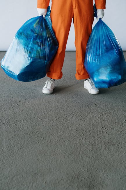 A person wearing orange work trousers and white sneakers is standing on a grey surface, holding two large blue plastic rubbish bags, one in each hand. The bags are filled with waste material, visible through the semi-transparent plastic, and appear to be tied at the top. The background is plain and neutral, emphasizing the focus on the individual and the rubbish bags. This scene illustrates waste collection or rubbish removal, which could be associated with private waste disposal services such as those provided by Waste Removal Catford, especially in contexts involving onsite clearance or alternative waste handling solutions outside of local authority collection. The setting suggests an indoor or a paved outdoor environment, with the individual actively transporting waste items, reflecting the practical aspect of rubbish removal tasks.