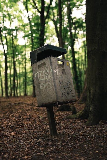A grey metal mailbox with graffiti markings on its surface, mounted on a black metal post that is leaning slightly to one side. The mailbox features a small, rectangular yellow sticker or writing area near the top and a slot for inserting mail. It has a black, slightly sloped lid covering the opening, which is partially open. The surrounding environment is a wooded area with tall trees and dense green foliage in the background, and the ground beneath the mailbox is covered with brown, uneven soil and scattered leaves. The natural light filters through the tree canopy, casting dappled illumination on the scene, creating a quiet, outdoor setting that subtly relates to the theme of private or independent waste handling, as the mailbox appears abandoned or weathered, possibly echoing the idea of off-site or alternative rubbish collection methods.
