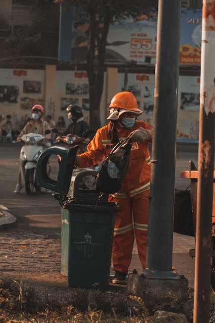 A person wearing orange work trousers and white sneakers is standing on a grey surface, holding two large blue plastic rubbish bags, one in each hand. The bags are filled with waste material, visible through the semi-transparent plastic, and appear to be tied at the top. The background is plain and neutral, emphasizing the focus on the individual and the rubbish bags. This scene illustrates waste collection or rubbish removal, which could be associated with private waste disposal services such as those provided by Waste Removal Catford, especially in contexts involving onsite clearance or alternative waste handling solutions outside of local authority collection. The setting suggests an indoor or a paved outdoor environment, with the individual actively transporting waste items, reflecting the practical aspect of rubbish removal tasks.