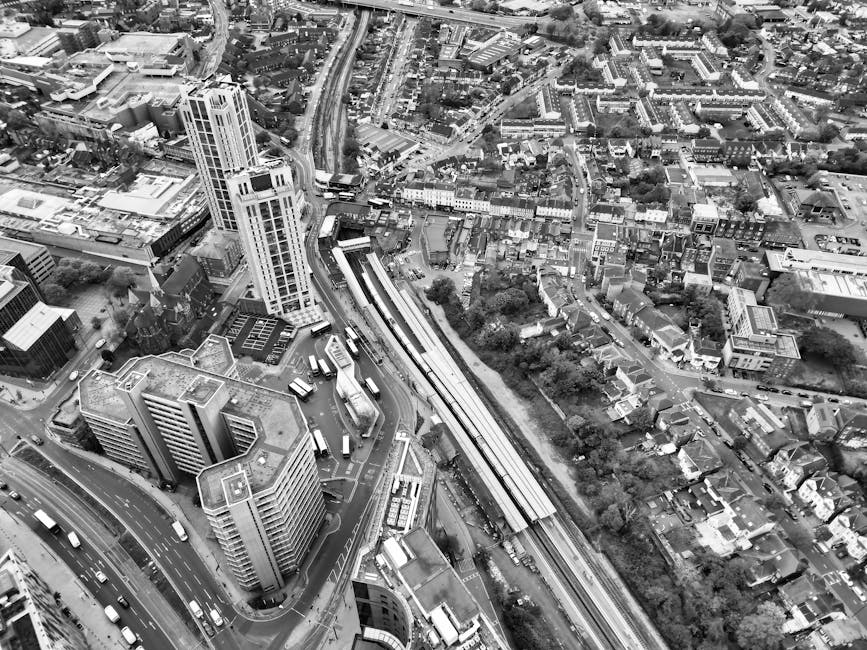 Aerial black-and-white view of a dense urban area featuring tall modern office buildings with glass facades and flat roofs in the foreground, alongside a multi-lane road with visible vehicles and buses. To the right, a railway line runs parallel, surrounded by a mix of residential houses and low-rise buildings, with tree-lined streets and small green spaces between structures. The background expands to include more streets, smaller buildings, and a network of roads and railways weaving through the urban landscape. The scene exhibits a combination of high-rise and traditional low-rise architectures, with textures of concrete, glass, and brick visible. The lighting appears natural, with diffused sunlight illuminating the scene evenly. This aerial image provides an opportunity to observe the layout of a city’s buildings, transport infrastructure, and residential areas, which can be relevant for services like independent waste collection and on-site clearance by companies such as Waste Removal Catford operating in the wider urban environment.