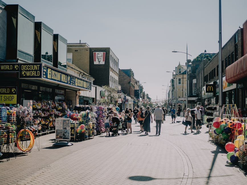 A wide pedestrian street in an urban shopping area is visible in the image, with various retail outlets lining both sides of the pavement. On the left, there is a store named 'Discount World' with a bright yellow sign and a display of colorful merchandise, including balloon bunches and toys, arranged outside on racks and carts. Adjacent to this, a sign for 'Kurt's Fashion' is partially visible, indicating other retail shops nearby. The right side features additional shops, including one with a dark awning and another with a bright yellow logo, possibly a fast-food restaurant like McDonald's, with outdoor displays and merchandise stands extending onto the pavement. Several pedestrians, some pushing strollers and carrying shopping bags, walk along the street, which is illuminated by natural daylight under a clear sky. The scene captures the lively, busy atmosphere of a shopping district with a mix of independent and chain stores, illustrating a typical setting where private waste removal services like Waste Removal Catford could assist with rubbish clearance during or after trading hours. The environment emphasizes urban commercial activity, with cluttered exterior display areas that might require rubbish disposal or street cleaning, aligning with local waste management needs.