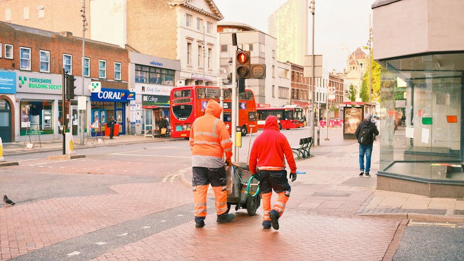 Two waste collection workers dressed in high-visibility orange and red safety clothing are pushing a wheeled bin on a busy city sidewalk. The worker in orange has their back to the camera, while the other, in red, is facing slightly sideways. The scene is set in an urban area with various storefronts visible in the background, including a building with a sign for YORKSHIRE BUILDING SOCIETY and another for CORAL betting shop. Red double-decker buses are present on the street, along with other pedestrians walking past, some near a bus stop and others along the pavement. The environment is well-lit with natural daylight, and the street features a mix of paved and brick surfaces, with a traffic light and street furniture such as benches and a glass display window on the right. This scene illustrates a typical scene of private rubbish collection or on-site waste clearance by Waste Removal Catford, serving local businesses and shops in the area.