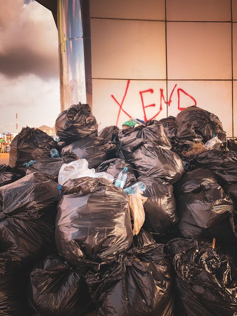 A large accumulation of black plastic rubbish bags is stacked against a tiled wall in an outdoor urban environment, with some bags appearing torn or loosely tied. Among the bags, a few discarded plastic water bottles and other small waste items are visible. The remnants of graffiti, including red spray-painted text that reads 'XEND,' are painted on the wall behind the bags. The scene is set in a paved area, possibly a service or loading zone, with a metallic column on the left side of the image. The lighting suggests late afternoon or early evening, with a cloudy sky casting a subdued, diffuse light over the scene. The overall impression is of an unmanaged waste accumulation that could be associated with private disposal or an alternative collection service, indicative of the need for professional rubbish removal, such as that provided by Waste Removal Catford.