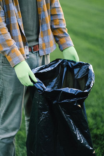 A person wearing a checked shirt with yellow, blue, and grey tones, alongside grey trousers and green gloves, is holding open a large black rubbish bag made of durable plastic material, with a metallic inner lining visible at the top. The individual is standing outdoors on a grassy area, with greenery and a blurred background suggesting a park or garden setting. The bag appears to be partially filled, and the scene is evenly lit, indicating daylight. This image relates to waste collection or rubbish removal activities, possibly involving joint or private waste handling services such as those provided by Waste Removal Catford, emphasizing proper on-site clearance of refuse in a community environment.