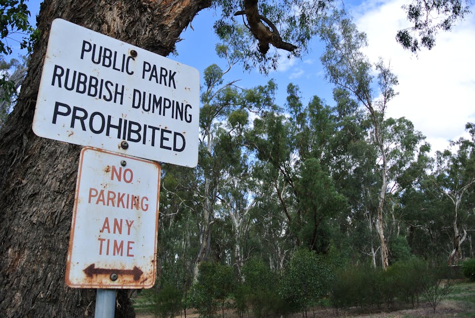 A white rectangular sign is mounted on a tree trunk in a natural outdoor setting, with the top portion displaying bold black text that reads 'PUBLIC PARK RUBBISH DUMPING PROHIBITED'. Below this, a smaller rectangular sign with rust markings states 'NO PARKING ANY TIME' in red capital letters and features a left-pointing arrow. The background consists of tall eucalyptus trees with slender branches and green foliage, set against a partly cloudy sky with patches of blue. The ground appears grassy and slightly overgrown, indicating a park or natural reserve. The signs are positioned close together, suggesting restrictions related to waste disposal and parking within the area, aligning with regulations that aim to prevent illegal dumping and vehicle obstruction in this natural environment. The overall scene emphasizes the importance of adhering to waste management rules in outdoor recreational spaces, with signs visibly indicating prohibited activities in this public park area managed under local regulations.