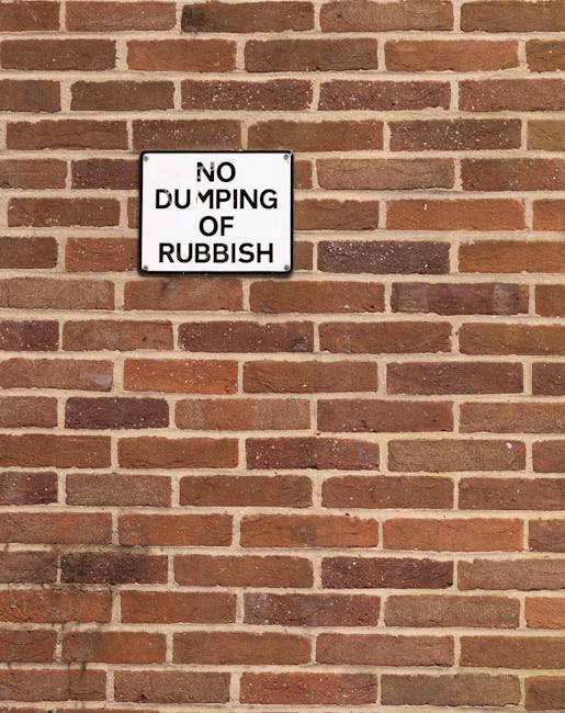 A white rectangular sign is mounted on a tree trunk in a natural outdoor setting, with the top portion displaying bold black text that reads 'PUBLIC PARK RUBBISH DUMPING PROHIBITED'. Below this, a smaller rectangular sign with rust markings states 'NO PARKING ANY TIME' in red capital letters and features a left-pointing arrow. The background consists of tall eucalyptus trees with slender branches and green foliage, set against a partly cloudy sky with patches of blue. The ground appears grassy and slightly overgrown, indicating a park or natural reserve. The signs are positioned close together, suggesting restrictions related to waste disposal and parking within the area, aligning with regulations that aim to prevent illegal dumping and vehicle obstruction in this natural environment. The overall scene emphasizes the importance of adhering to waste management rules in outdoor recreational spaces, with signs visibly indicating prohibited activities in this public park area managed under local regulations.