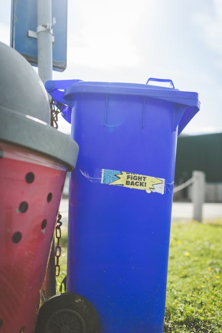 A tall, rectangular blue plastic rubbish bin with a closed lid, positioned outdoors on a grassy area. The bin has a yellow sticker with the words 'FIGHT BACK!' on its front. To the left of the blue bin, part of a red waste container with black holes and a grey lid is visible, attached to a black wheel and chain mechanism. In the background, there is a light blue sign mounted on a pole, a grey fence, and a cloudy sky, suggesting a suburban or urban environment. The scene appears to be part of a private waste collection site, with a focus on managing rubbish efficiently in a setting where space may be limited, such as tight access areas. The natural daylight highlights the textures of the plastic bins and the surrounding environment, aligning with waste removal services that provide on-site clearance solutions for various disposal needs.